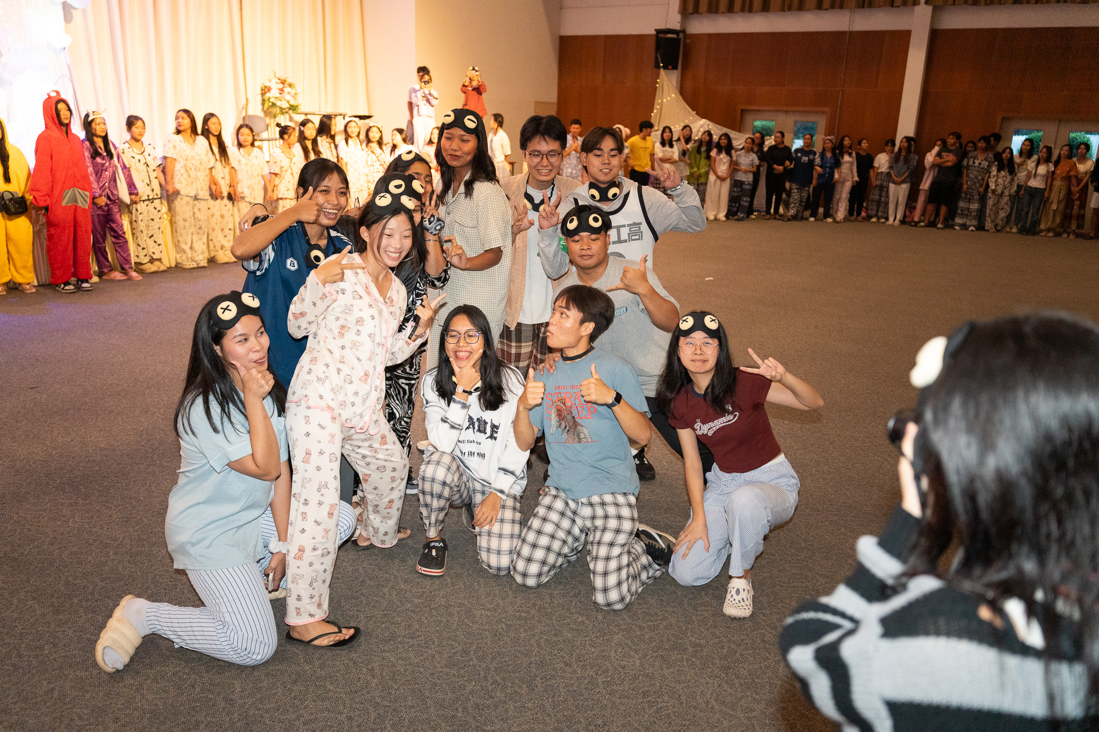 Energetic students dressed in sleepwear for a pajama-themed party give thumbs-up and peace signs in a large hall filled with their peers.